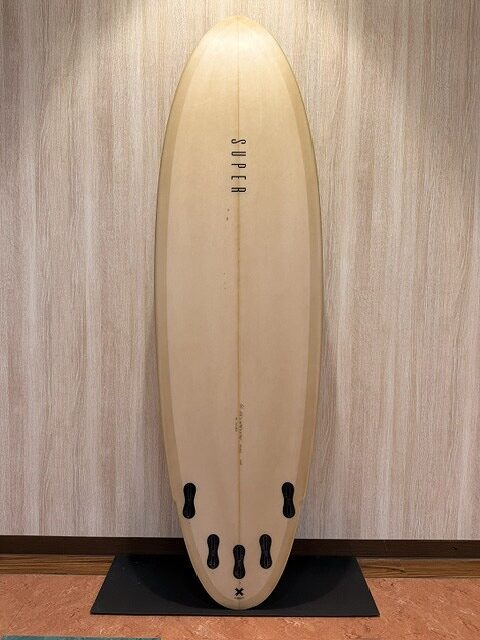Beige surfboard standing upright against a wall on a black display mat, indoors.