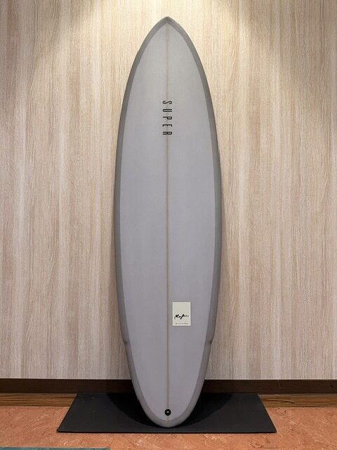 Gray surfboard with black edge, upright against a wood-paneled wall on a black mat indoors.