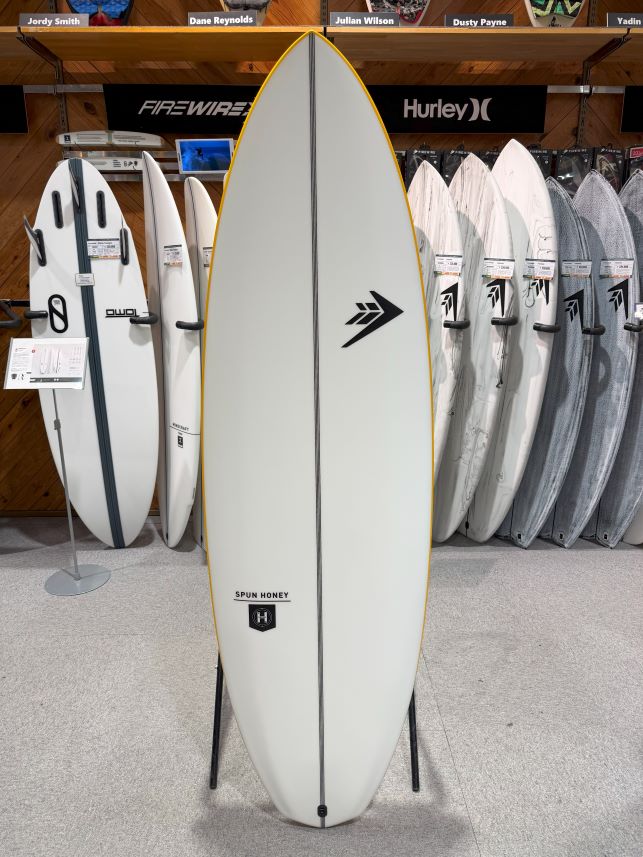 White surfboard with a yellow-edged rail on a stand in a showroom, surrounded by other boards and brand banners.