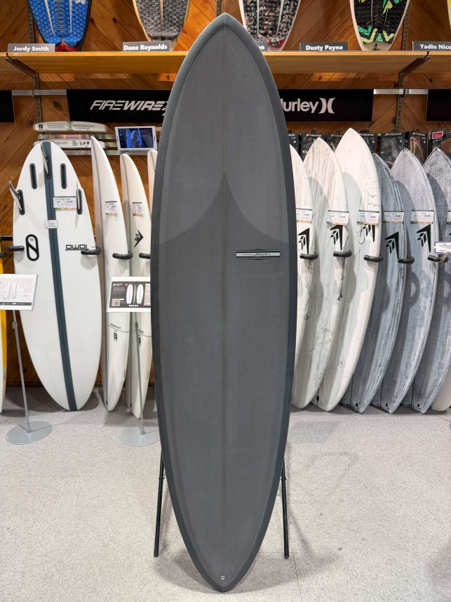 Large gray surfboard on a stand in a surf shop, with white and patterned boards lining the wall behind.