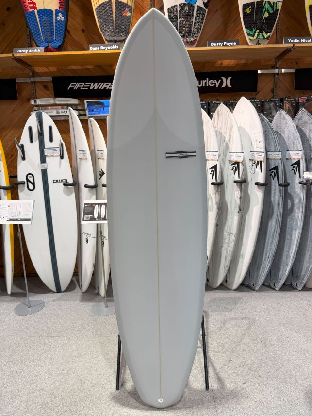 Tall light-gray surfboard on a stand in a shop, surrounded by other boards and racks in the background.
