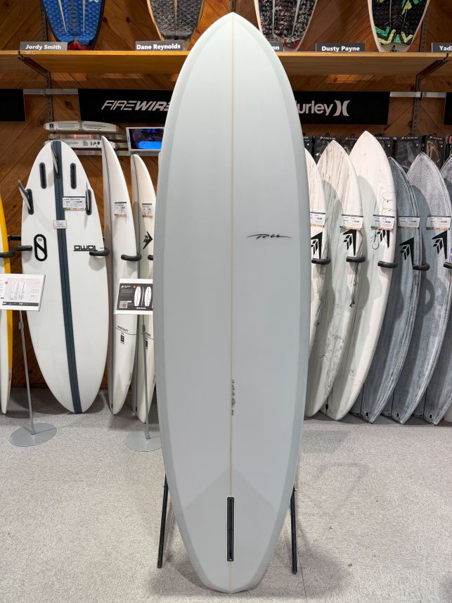 Tall white surfboard standing upright on a stand in a shop, surrounded by other boards on racks.