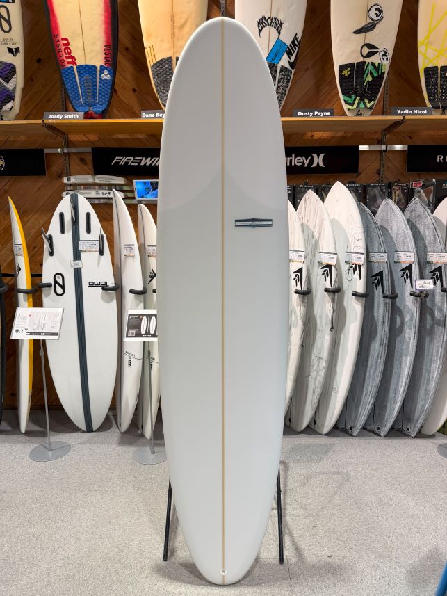 Tall white surfboard on display stand in a shop, surrounded by other boards on racks and wall shelves.