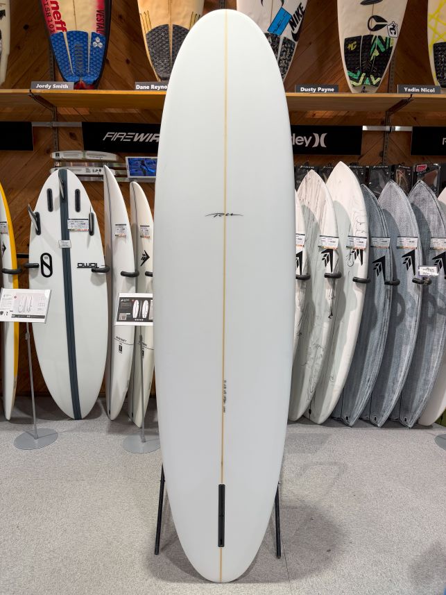White surfboard displayed upright on a stand in a store, featuring a single beige stringer down the center and surrounded by other boards on racks.