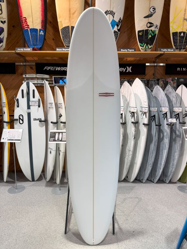 White surfing board on a stand in a shop, surrounded by other boards on racks and on the wall displays behind it.