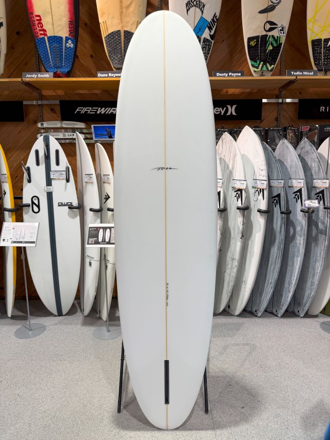 White surfboard on a stand in a shop, centered among other boards, with a narrow wood stringer running lengthwise and a small fin box near the tail.