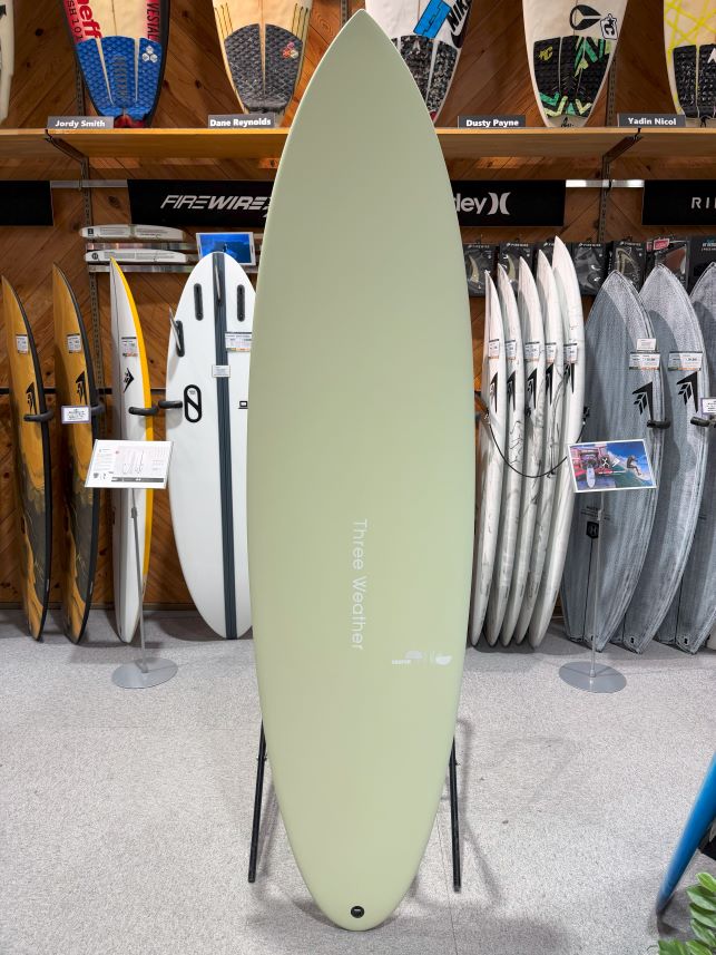 Pale green surfboard standing upright on a black stand in a surf shop, with racks of other boards and paddles visible on the walls behind it.