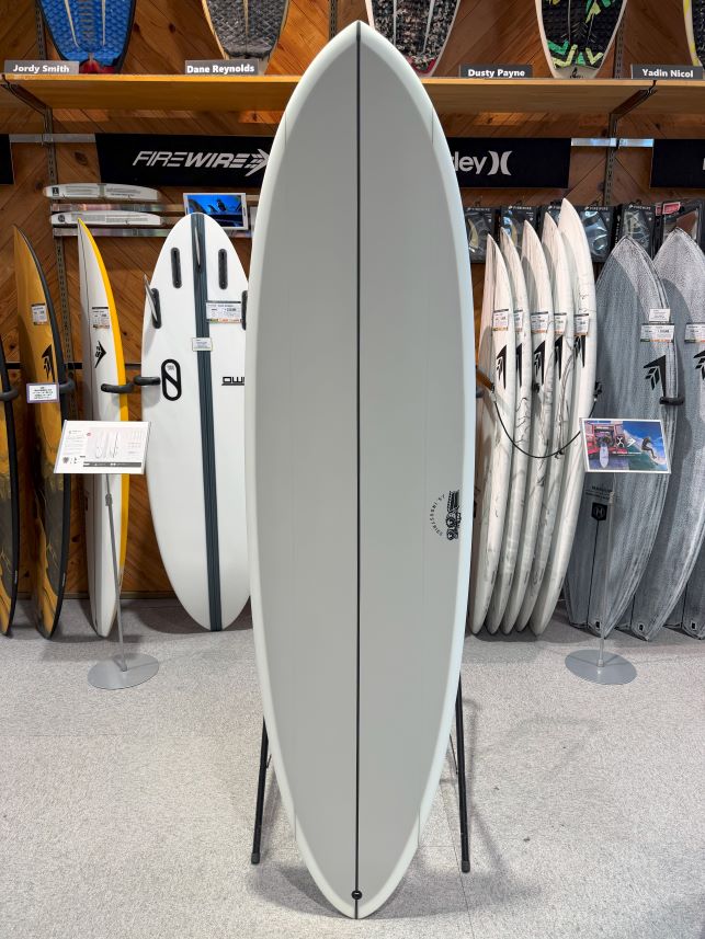 Tall gray surfboard on a display stand in a surf shop, with other boards on racks behind it.