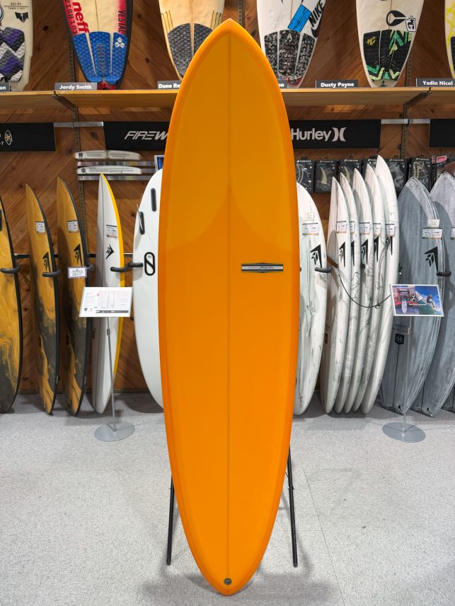 Orange surfboard standing upright on a display stand in a surf shop, with many other boards on racks in the background.