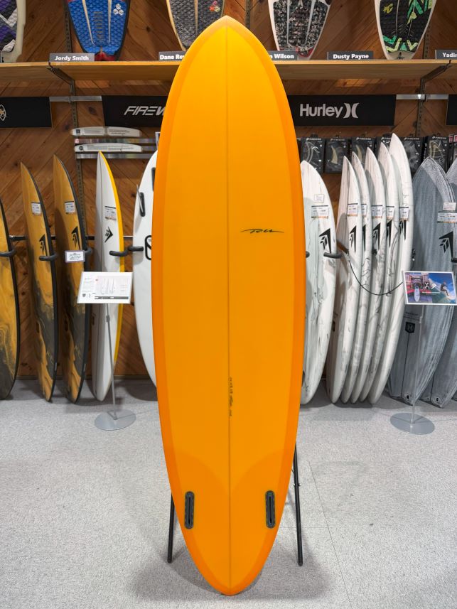 Bright orange shortboard standing upright on a display stand in a surf shop, with other boards visible in the background.