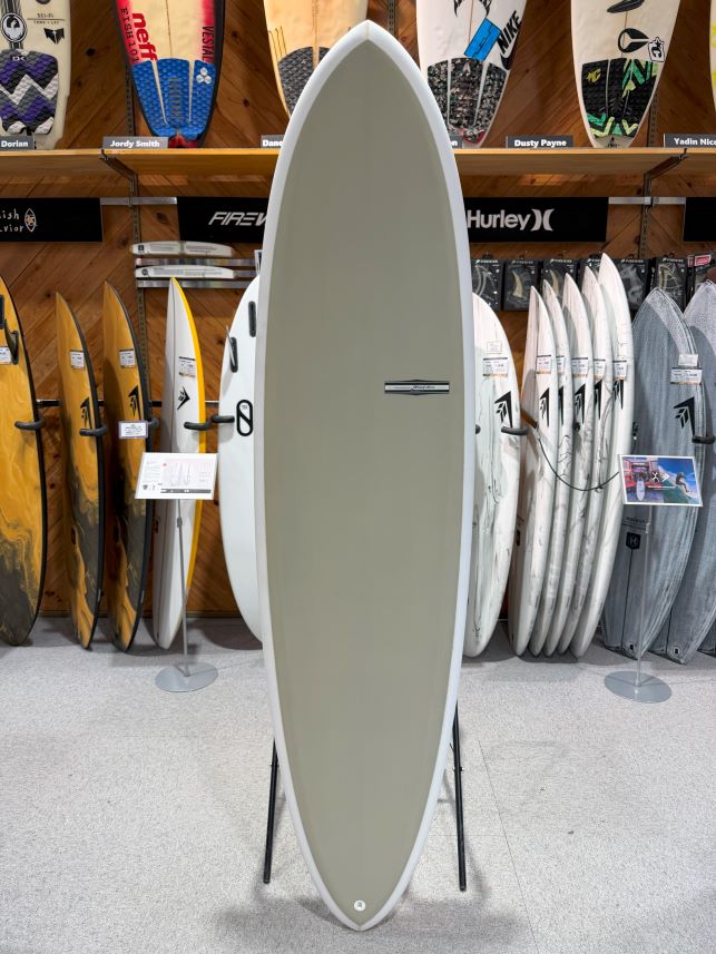 Tall gray surfboard on a stand in a surf shop, with colorful boards displayed behind it on racks
