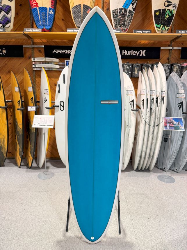Blue surfboard on a stand in a shop, surrounded by assorted surfboards on racks behind it.