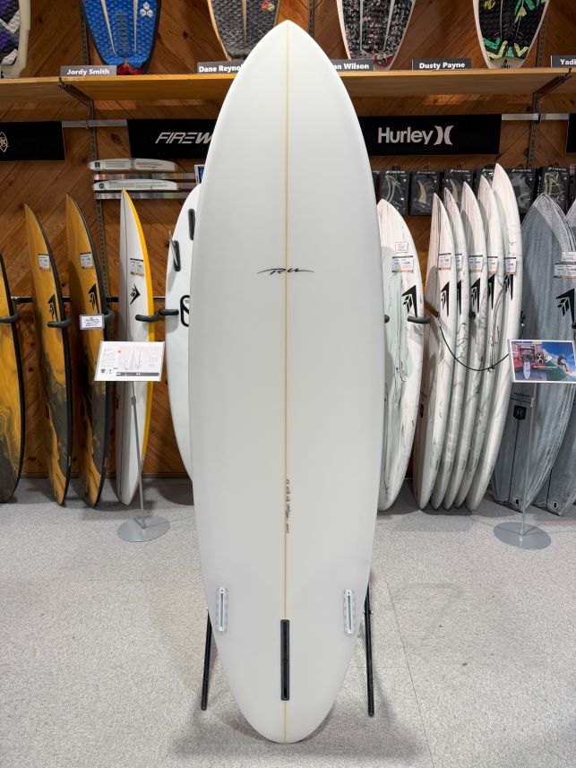 White surfboard on a display stand in a shop, with other boards on racks behind it.