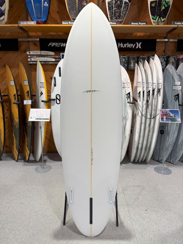 White shortboard on a display stand in a surf shop, surrounded by other boards on racks behind it.