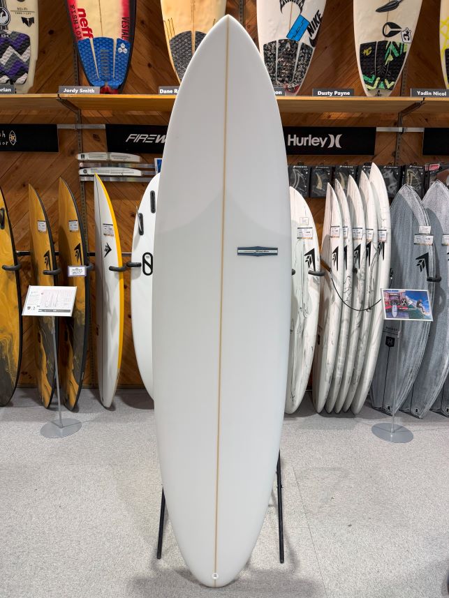 White shortboard surfboard on a display stand in a surf shop, with other boards visible in the background.