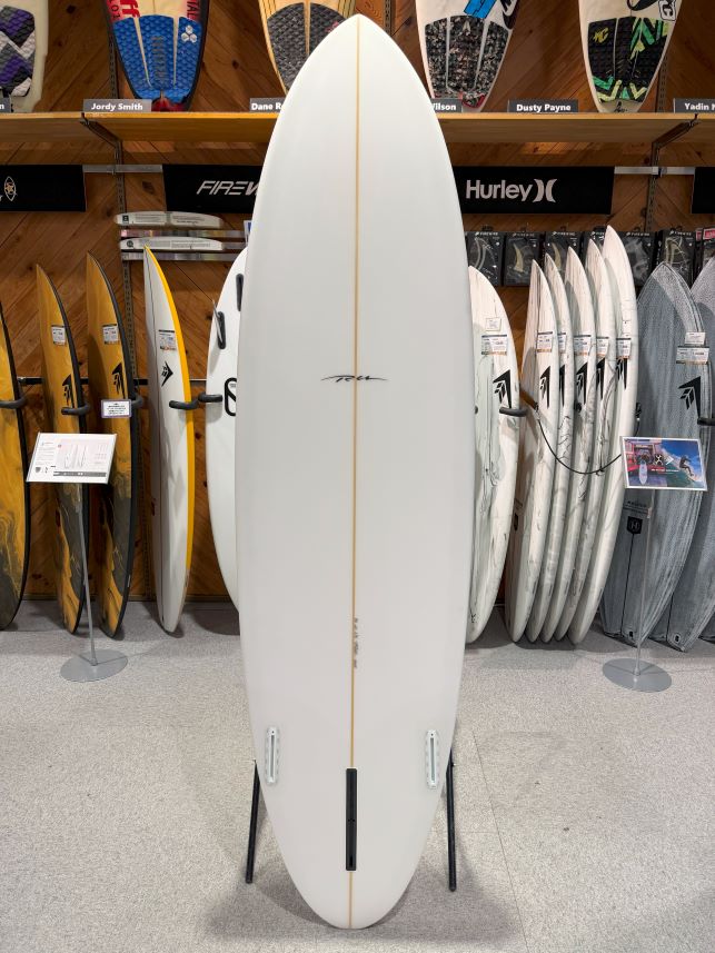 White surfboard on a display stand in a shop, centered with other boards on racks behind.