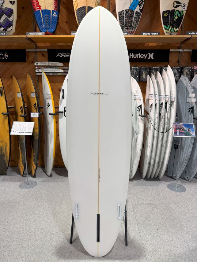 White surfboard with a thin gold stringer on a display stand in a surf shop, surrounded by other boards