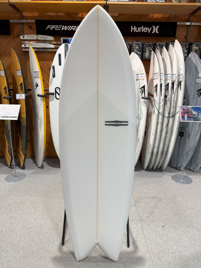 White surfboard displayed upright on a rack in a surf shop, surrounded by other boards and signage.