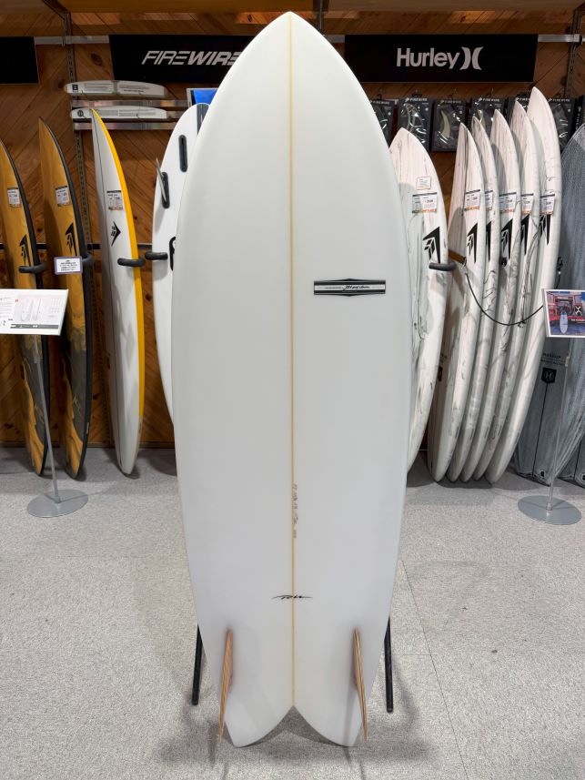 White surfboard on a display stand in a surf shop, with other boards lined up behind it on wooden shelving.
