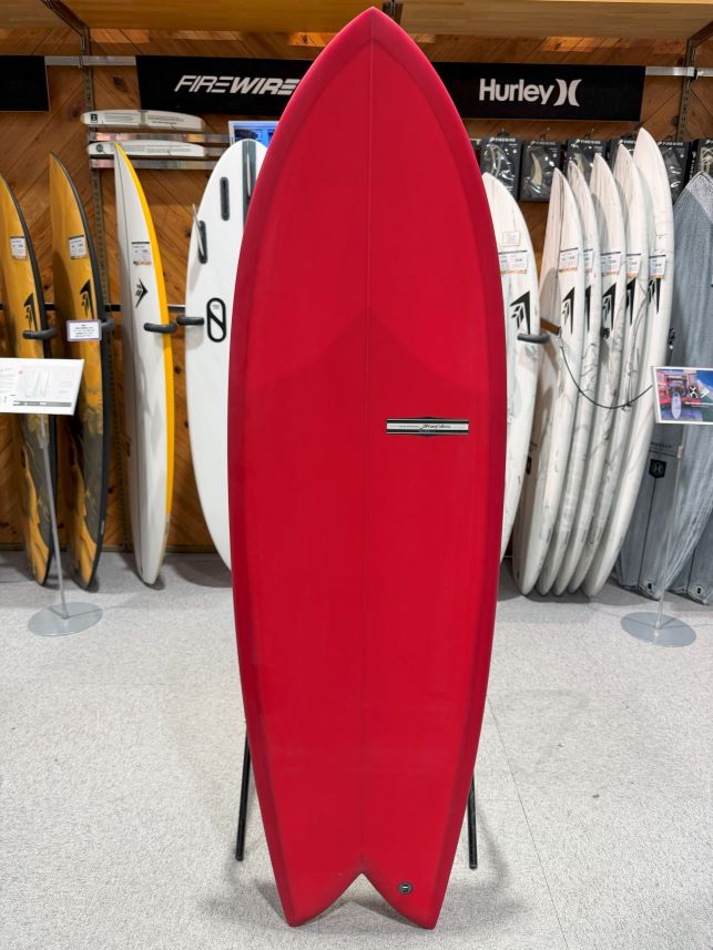 Red surfboard standing upright on a display rack in a surf shop, with other boards and brand banners visible in the background.