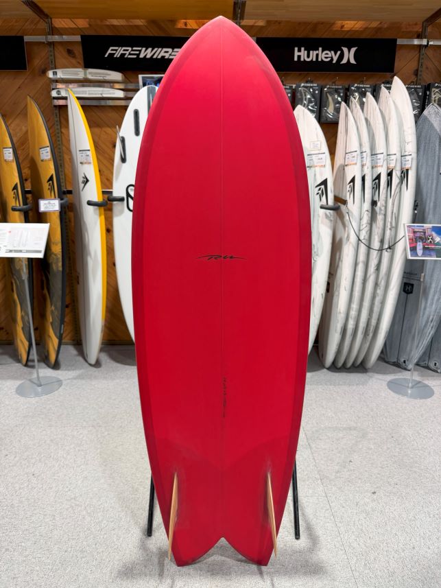 Red surfboard on display in a surf shop with other boards visible in the background.