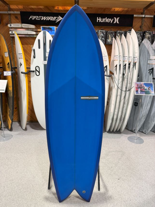 Blue shortboard surfboard on a stand in a store, surrounded by other boards and banners in the background.