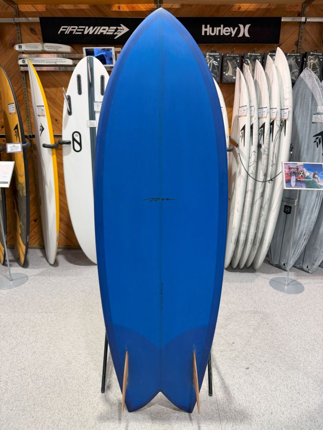 Blue surfboard on a stand in a surf shop, with white and patterned boards on racks in the background.