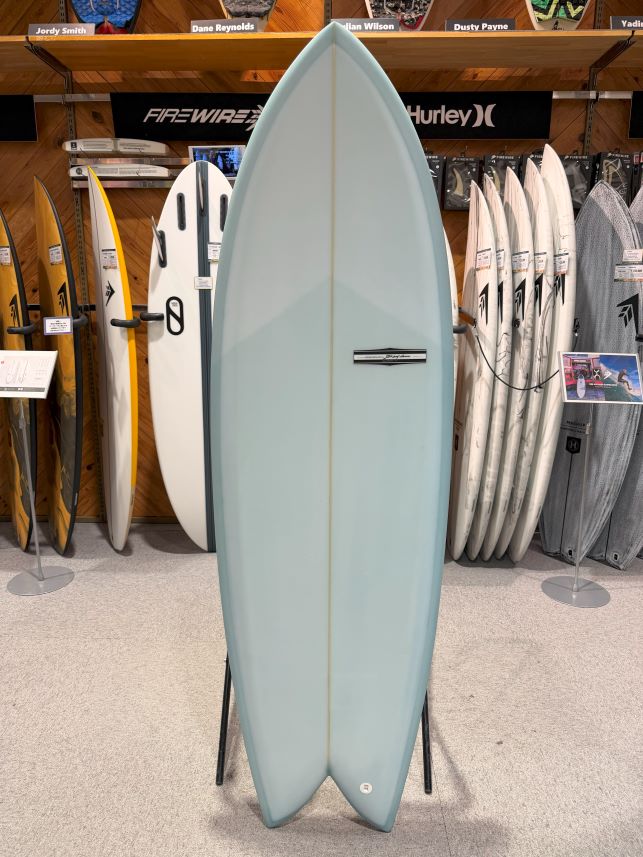 Light blue surfboard on a display stand in a surf shop, surrounded by other boards and shelves.
