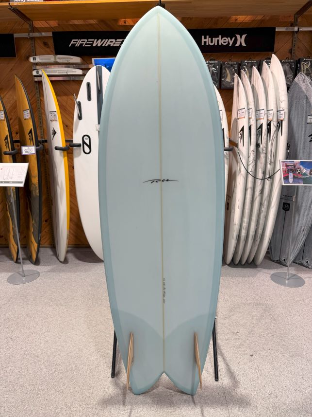 Light blue longboard surfboard on a display stand in a surf shop, with other boards and brands like Firewire and Hurley in the background.