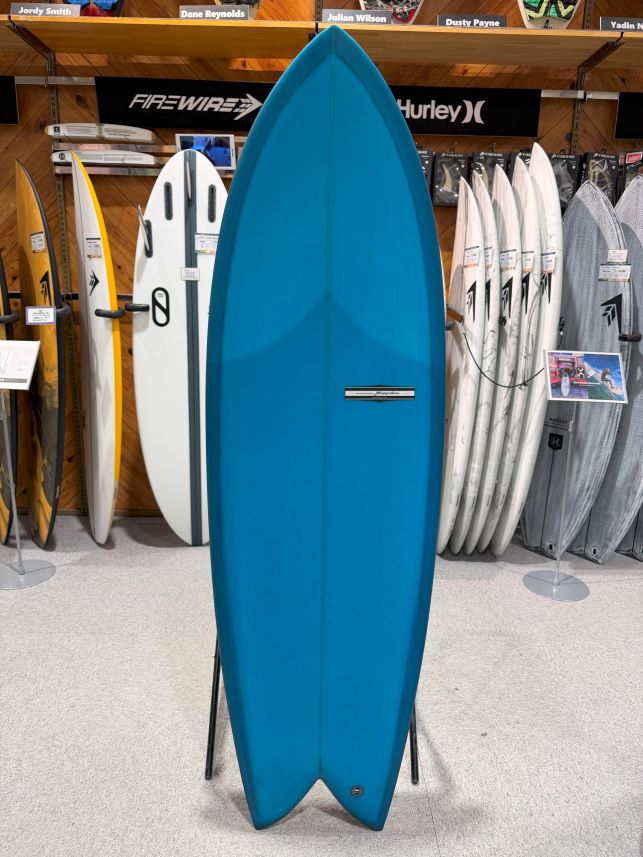 Tall blue surfboard on a display stand in a surf shop, with other boards lined up behind it.