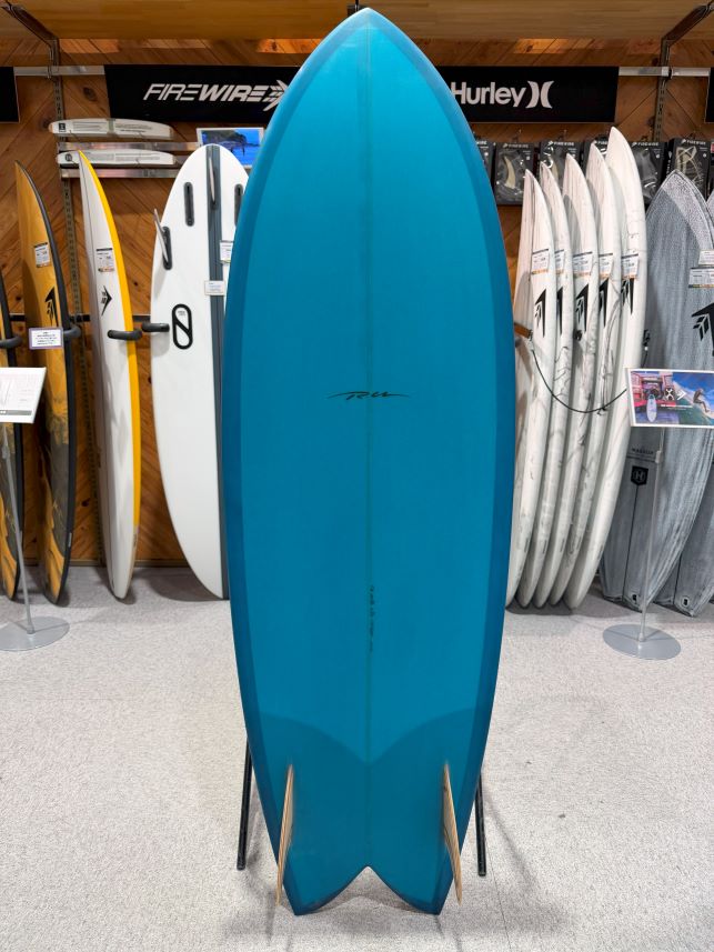 Blue surfboard standing upright on a display rack in a surf shop, with other boards and brand signage in the background.