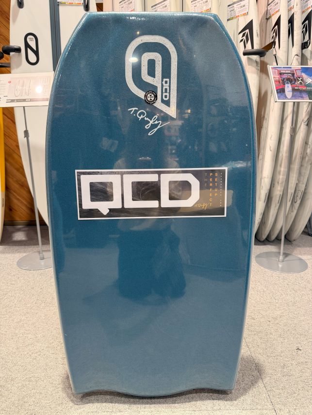 Blue stand-up surfboard with white 'QCD' logo, displayed upright in a store.