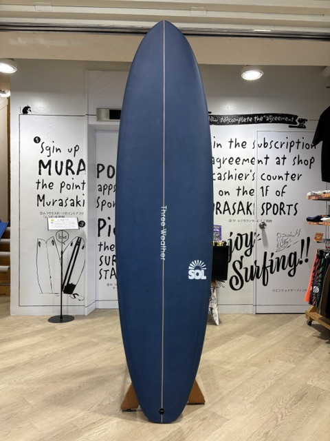 IMG_9749 - SHOP NEWS | ムラサキスポーツオンラインストア 通販 Blue surfboard on a wooden stand inside a shop, centered against a wall with black-and-white graphic text and posters.