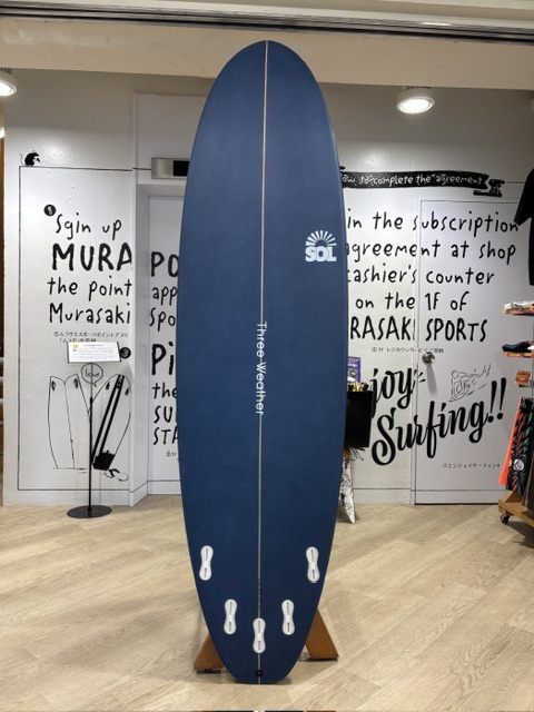 IMG_9750 - SHOP NEWS | ムラサキスポーツオンラインストア 通販 Blue surfboard on display in a shop, with a white center stringer and SOL logo on the upper half.