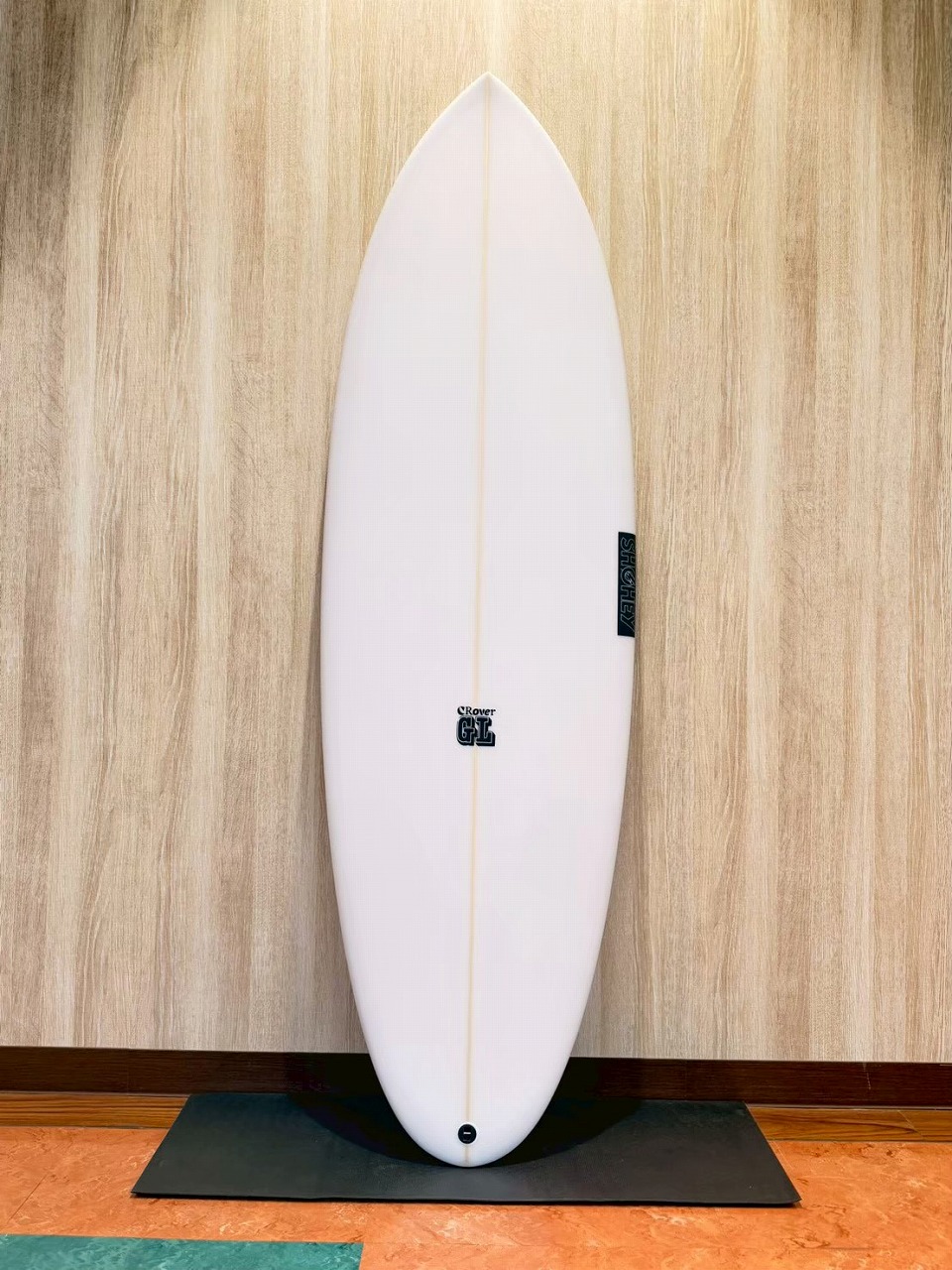 White surfboard standing upright on a black mat against a wood-patterned wall, showing a small logo near the center and a vertical tag on the right edge.