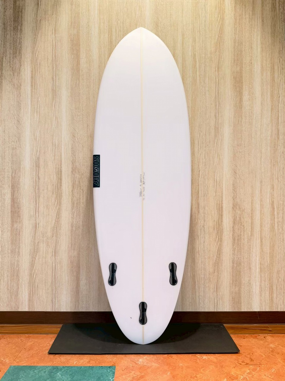 White surfboard standing upright against a wood wall on a black mat, three fin boxes visible on the bottom half.