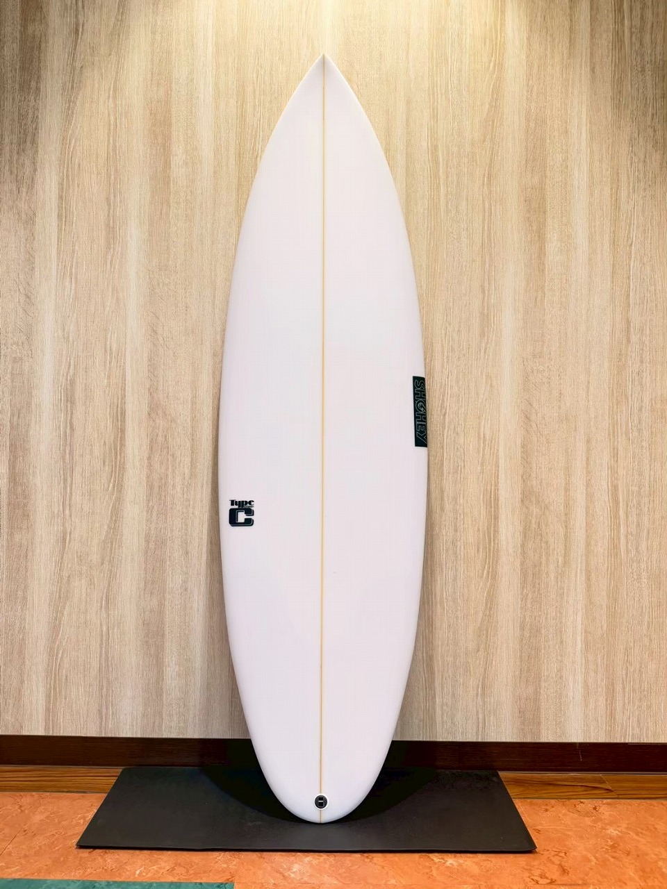 White surfboard standing upright against a wood-paneled wall on a black mat indoors, showing a central stringer and tail logo.