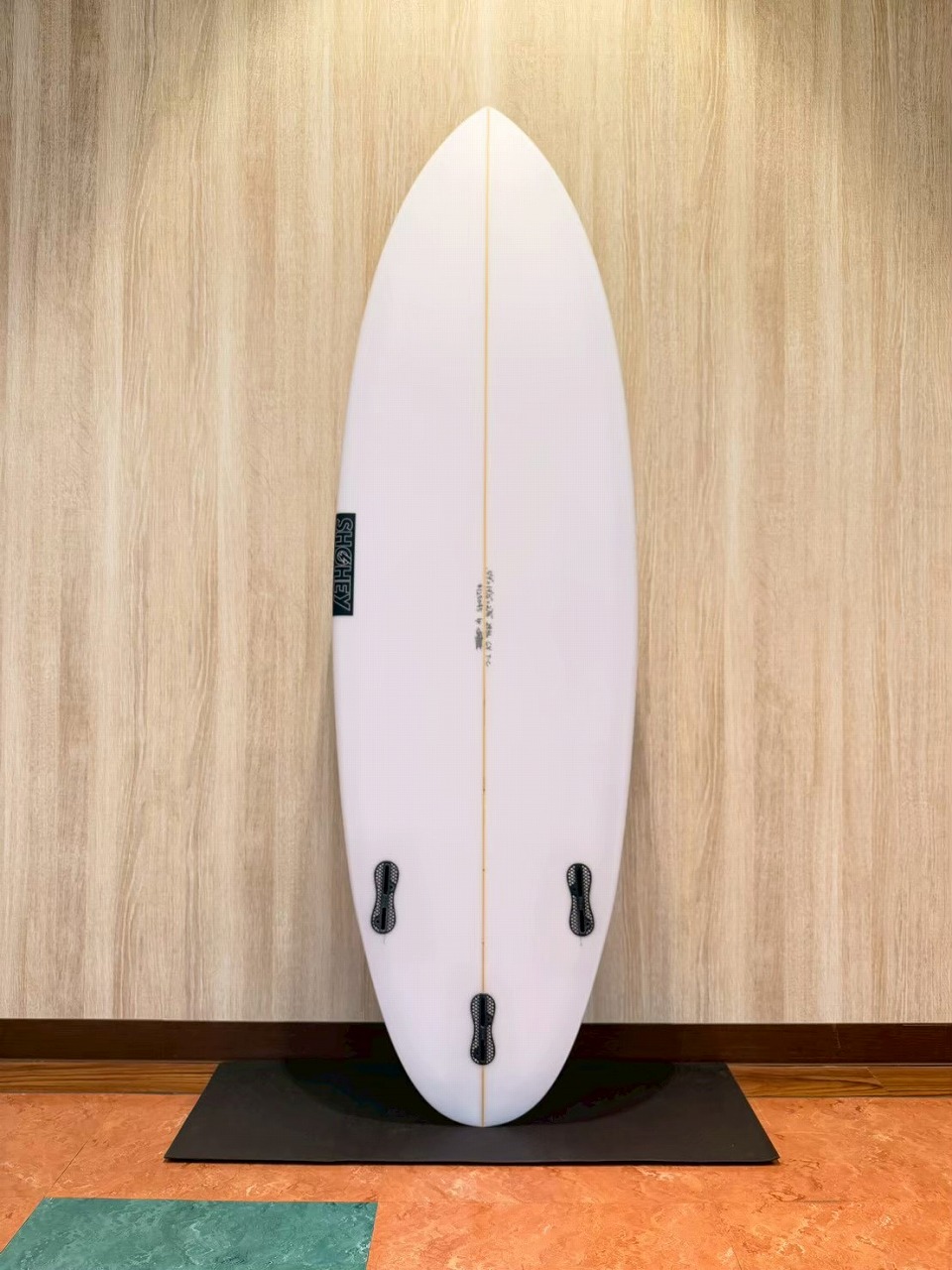 White surfboard standing upright indoors against a wood wall on a black mat, with an orange center string and four fin boxes on the bottom half.