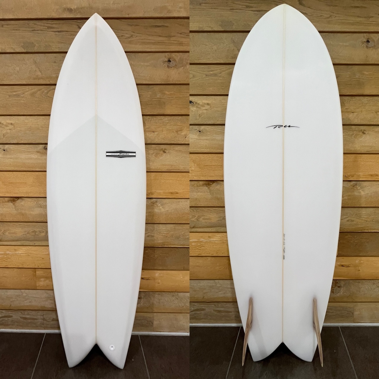 Front and back views of a white shortboard propped against a wood wall; a small rectangular badge is on the front.