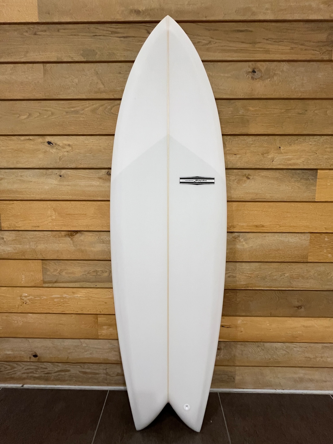 White surfboard standing upright against a wooden wall, with a central stringer and small logo plate.