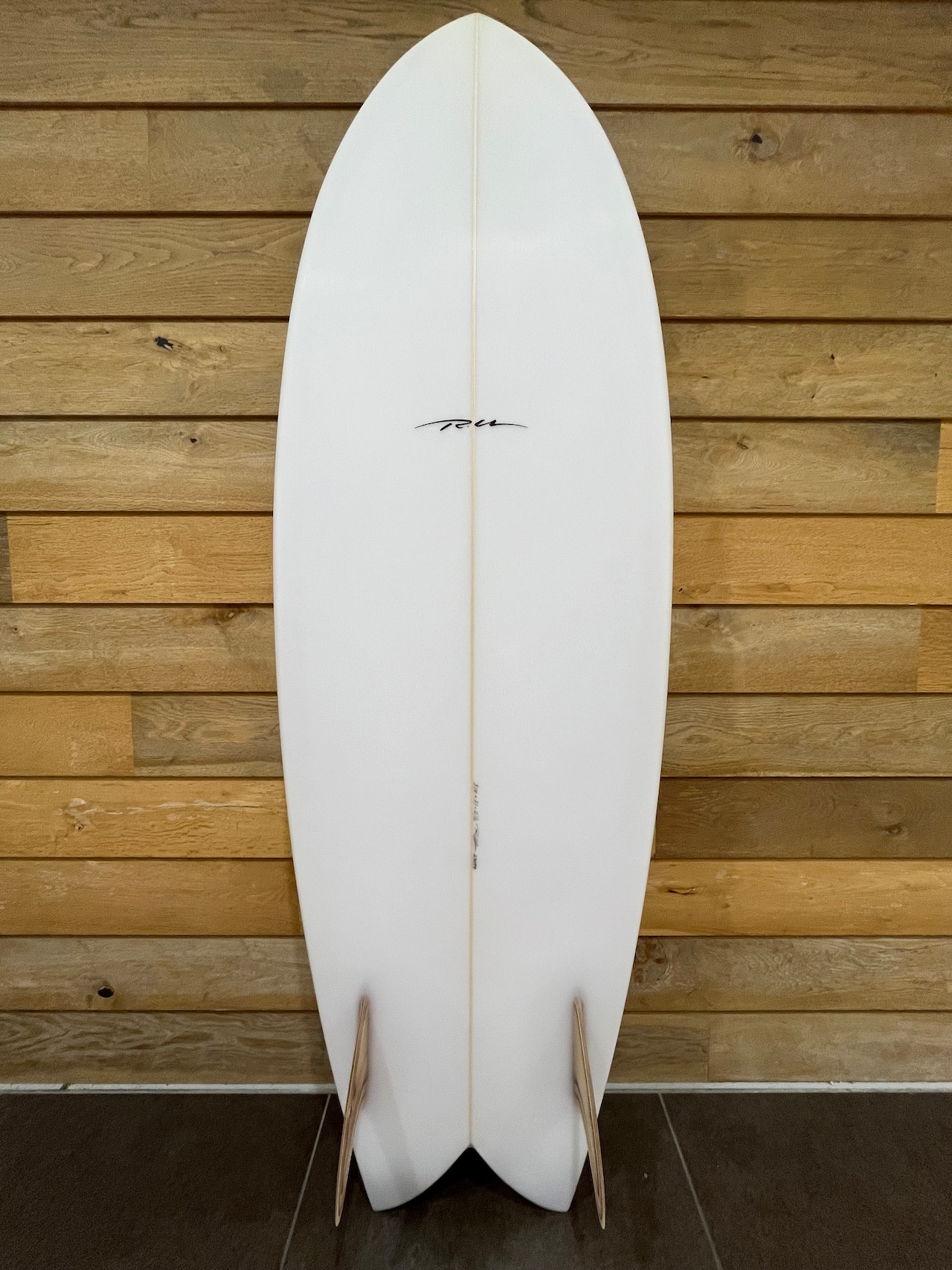 White surfboard standing upright against a wooden panel wall, with two light-colored fins at the tail end.