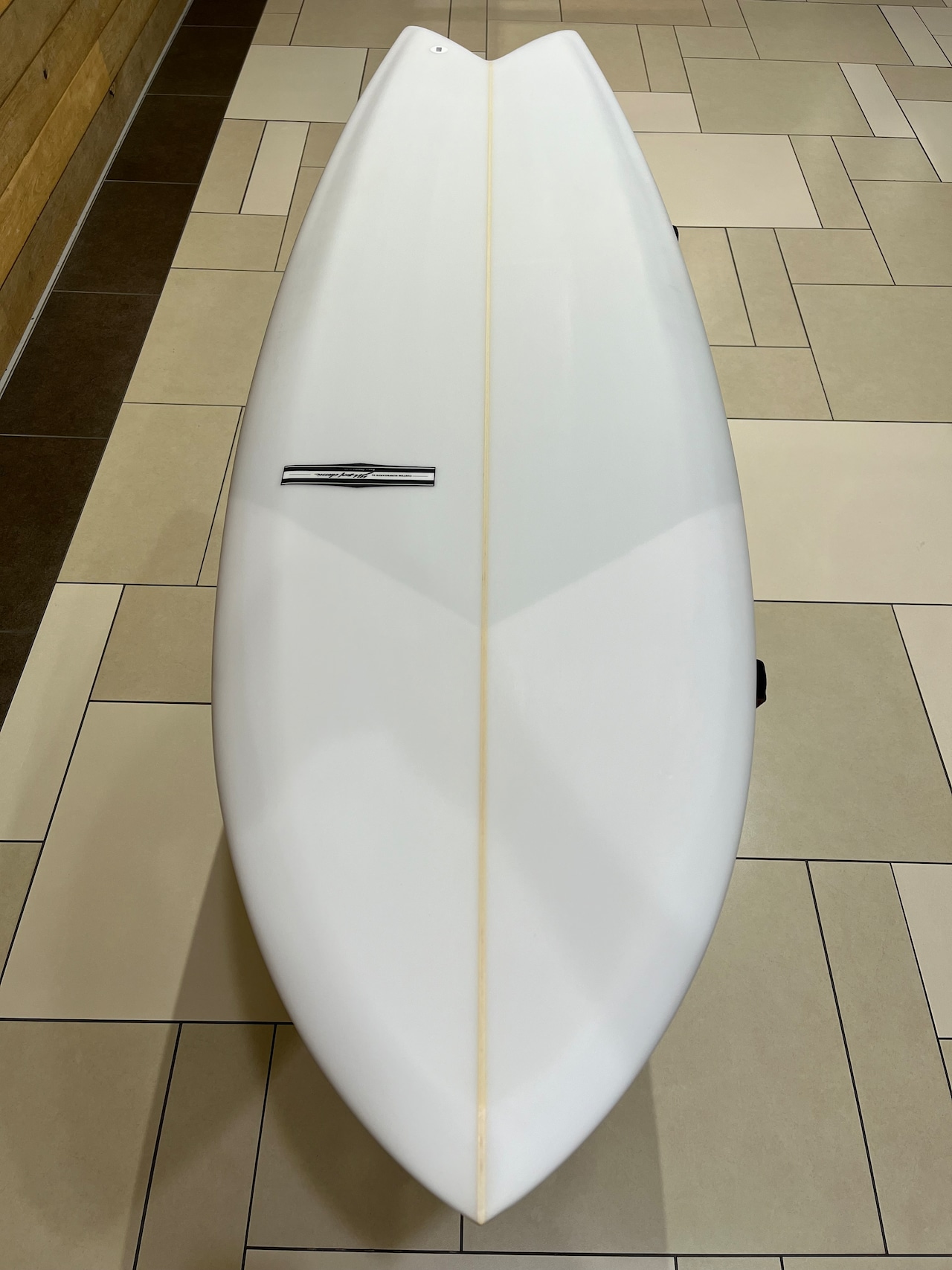 White surfboard viewed from above, lying on a beige tiled floor with a pale center stringer and a small black-and-white brand badge on the left side.