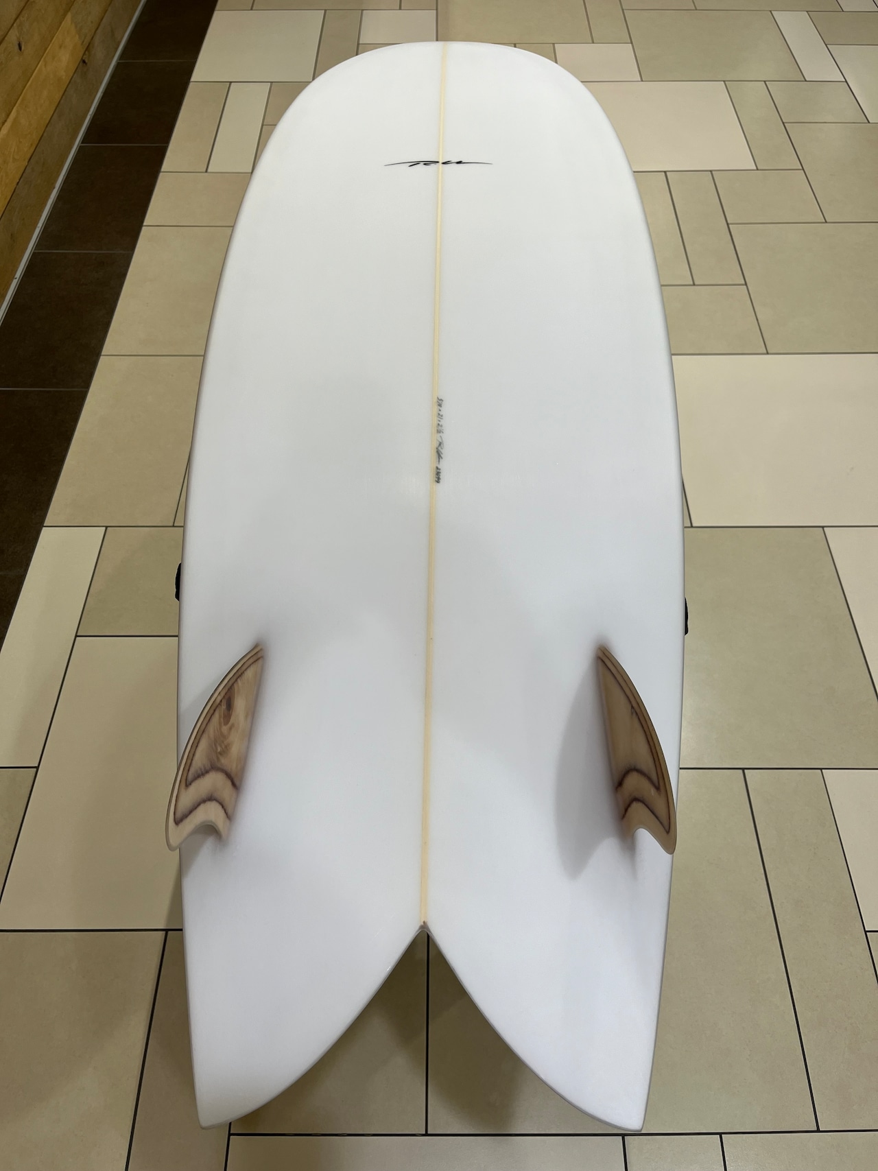 Top-down view of a white surfboard with a beige stringer and two wooden fins near the tail, resting on a tiled floor.