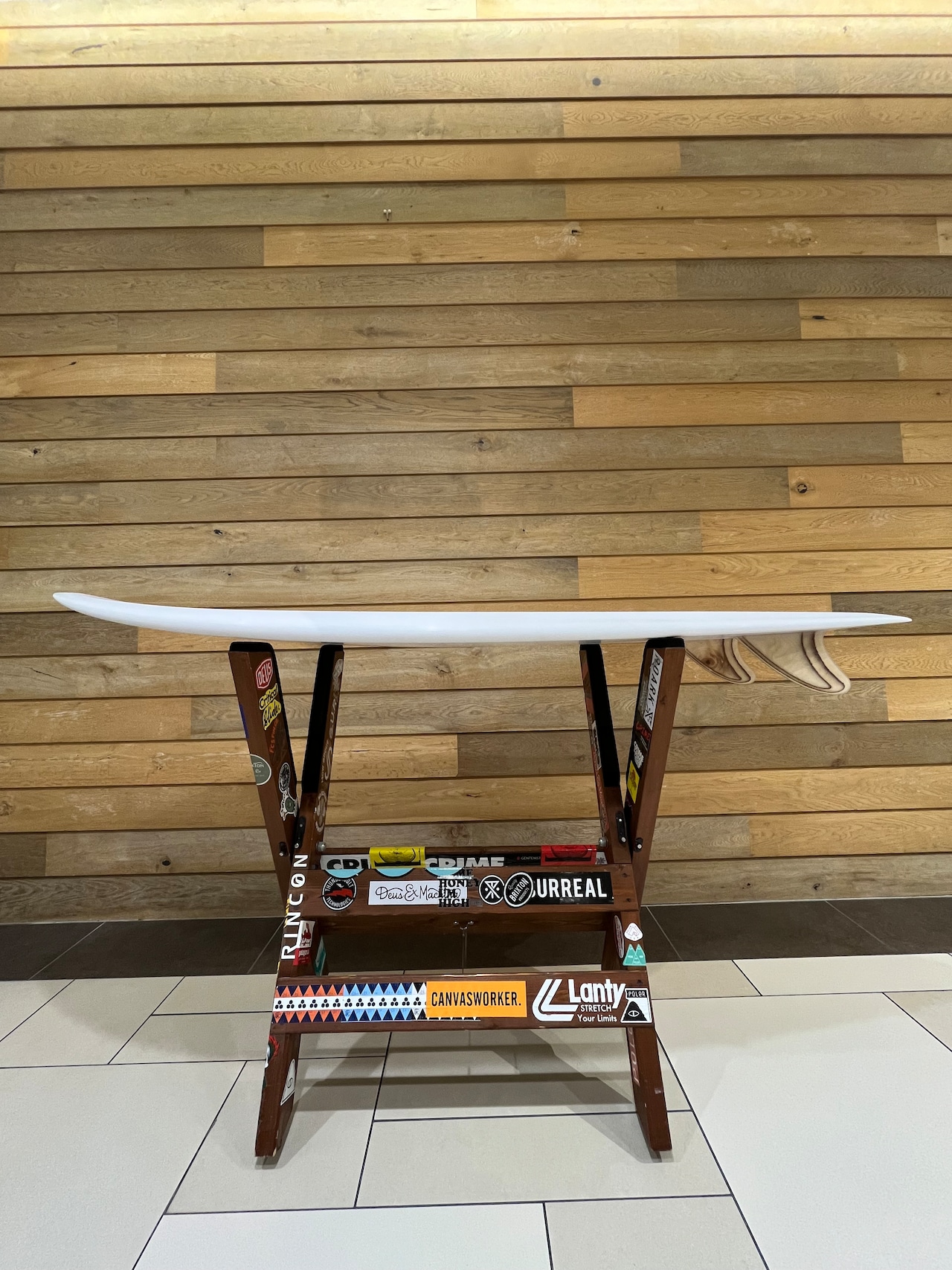 White surfboard resting on a sticker-covered wooden rack indoors against a light wood-paneled wall.