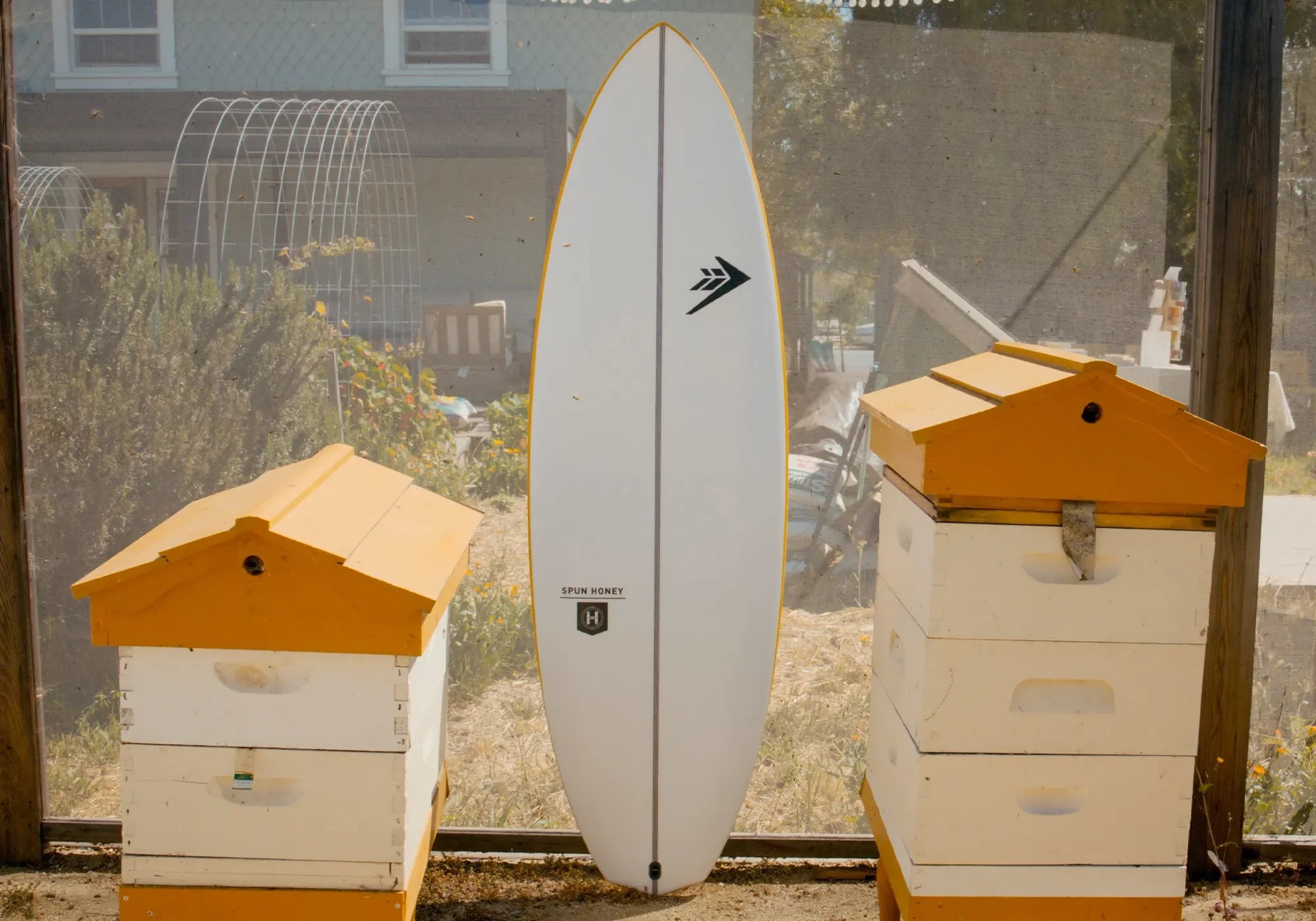 White surfboard with a black chevron logo stands upright between two yellow beehives in a sunny backyard.