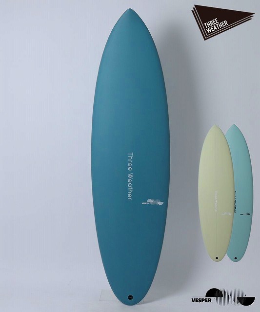 Tall blue surfboard with Three Weather branding stands against a pale wall, beside two smaller pastel boards (yellow and light blue).