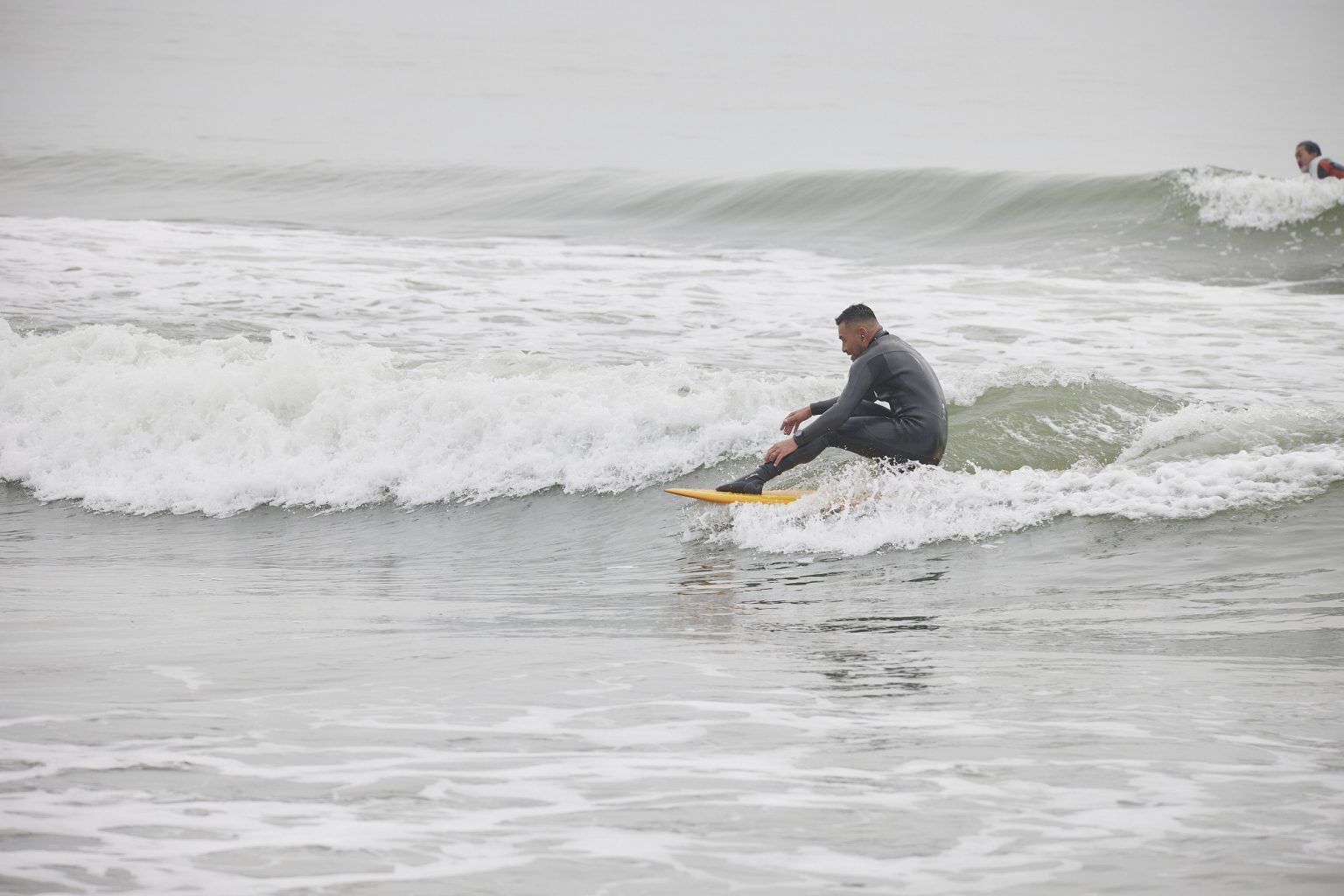 Surfer in a gray wetsuit riding a small wave on a yellow board; another surfer visible in the background.