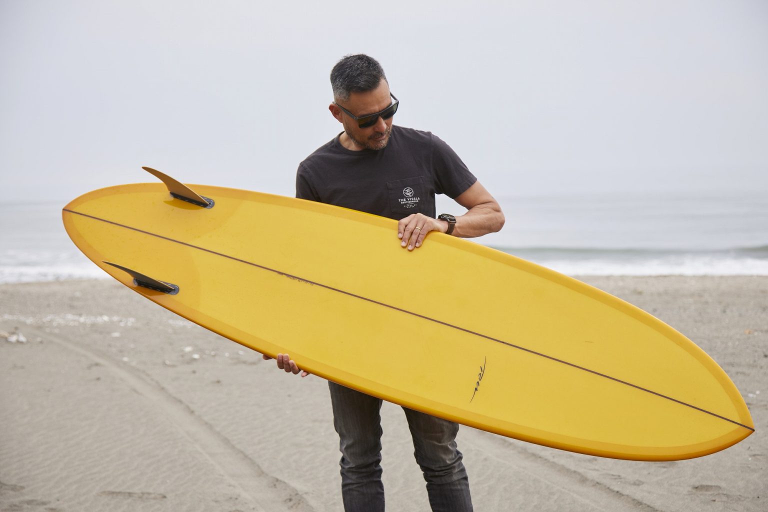 Man on a beach holding a bright yellow surfboard across his chest, wearing sunglasses.