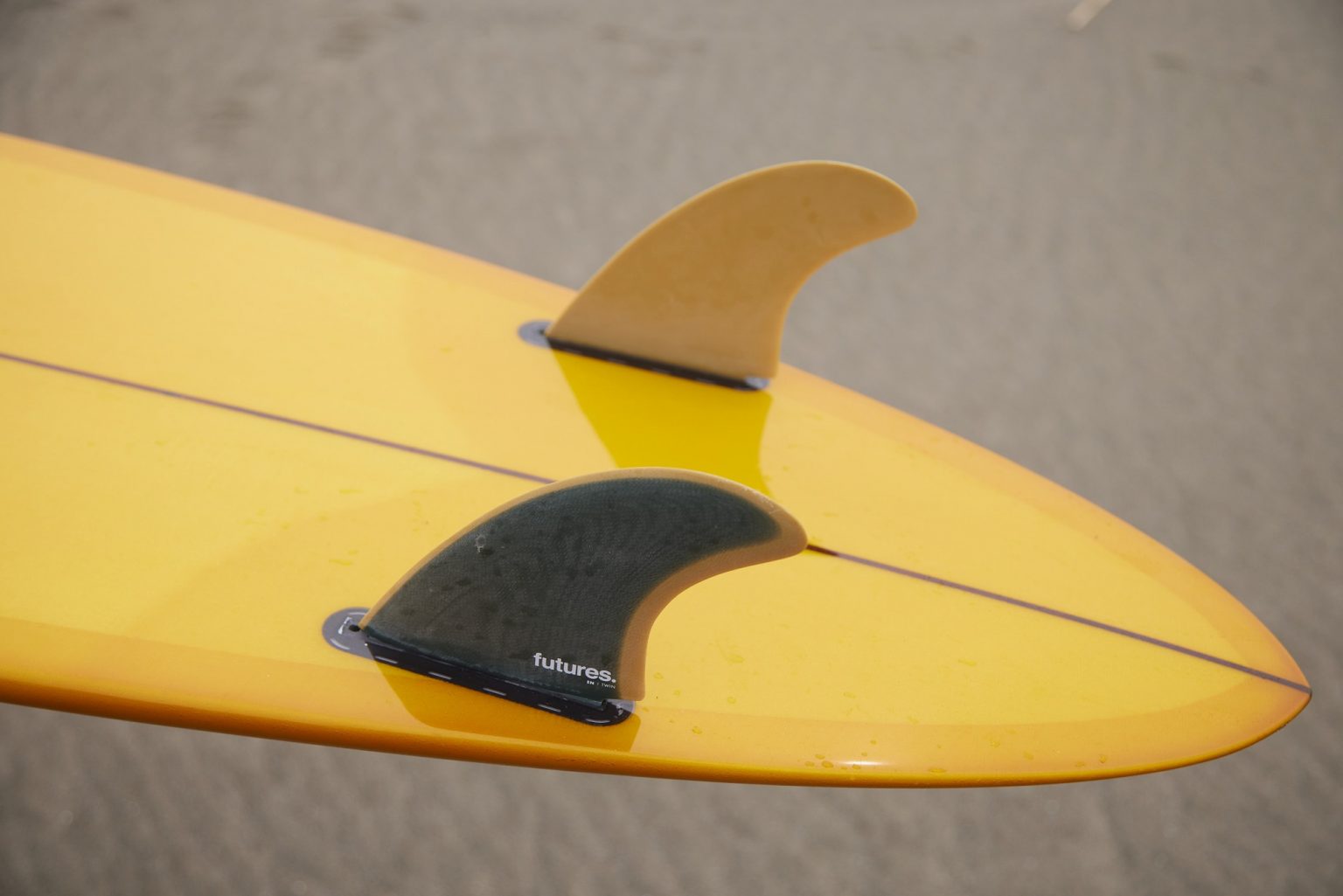 Close-up of a yellow surfboard tail with two fins: a black Futures fin and a smaller beige fin