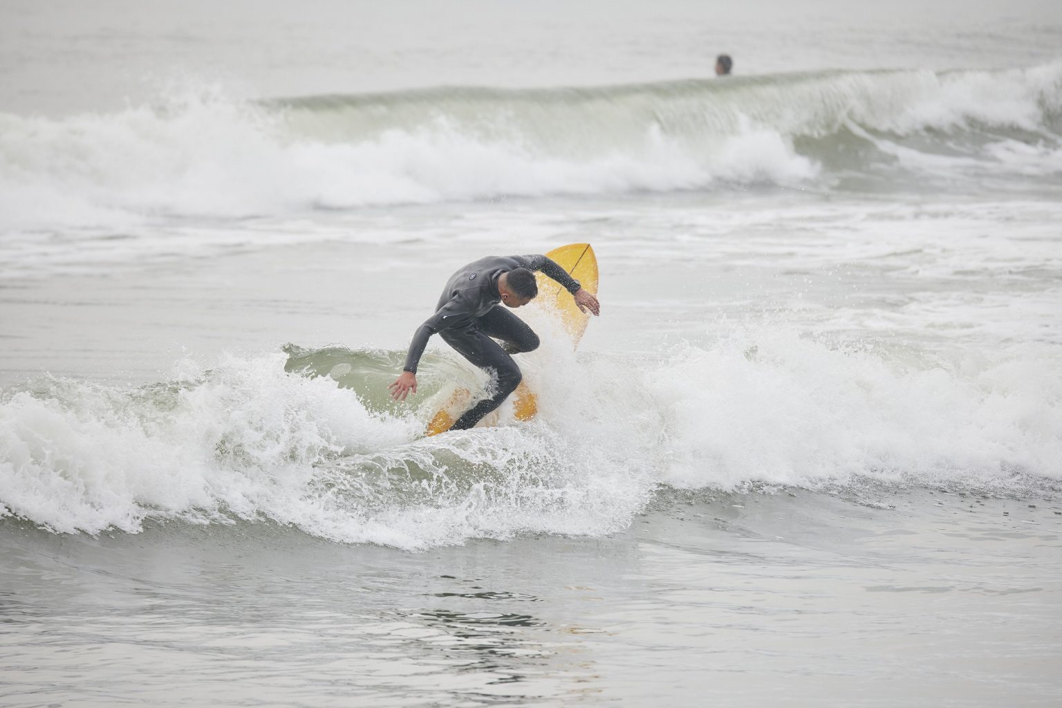 Surfer in a gray wetsuit rides a yellow surfboard on a small, frothy wave at the beach.